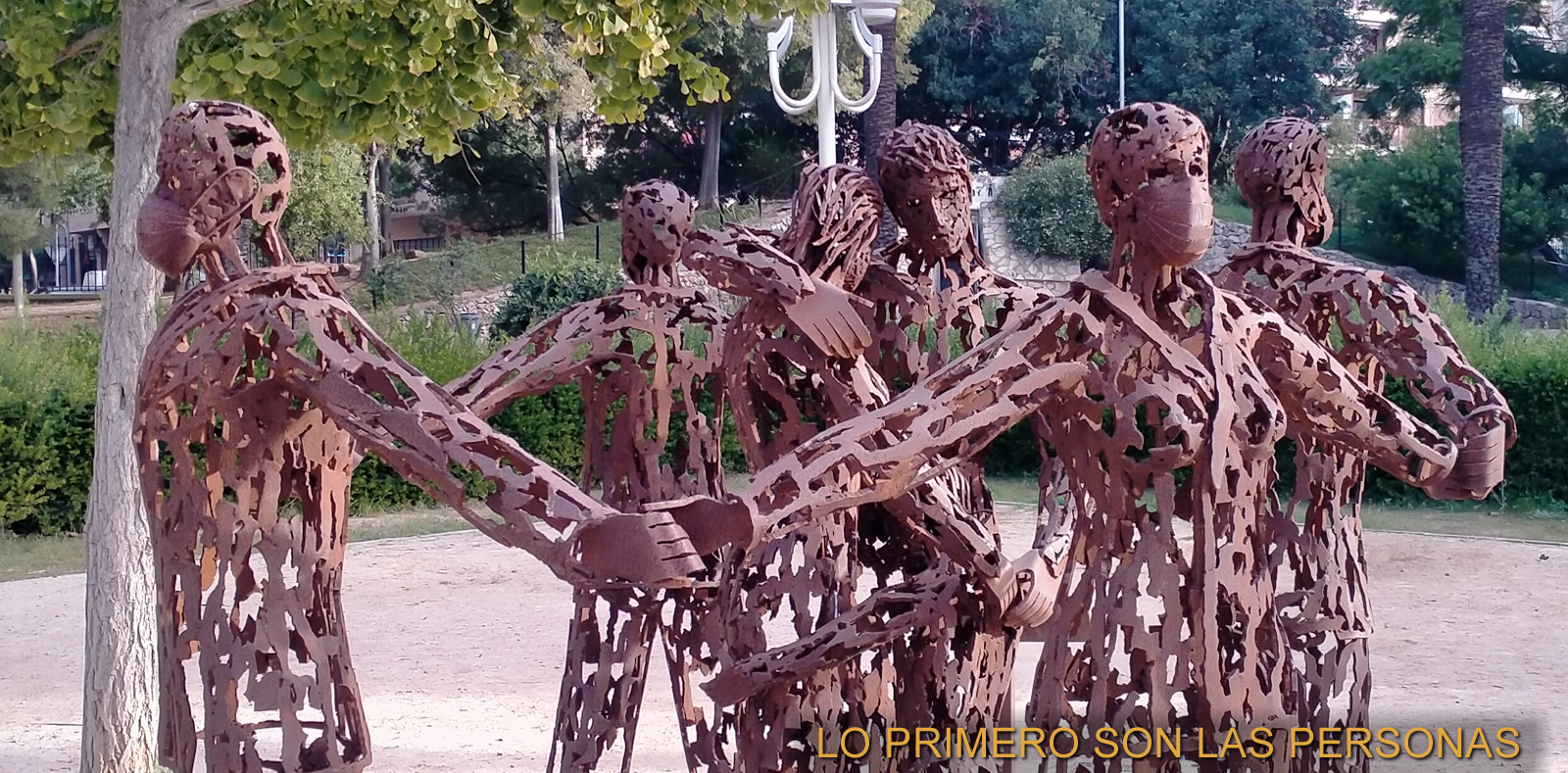 Escultura de figuras humanas de hierro cogidas de la mano formando un corro en un parque, con el lema «Lo primero son las personas»